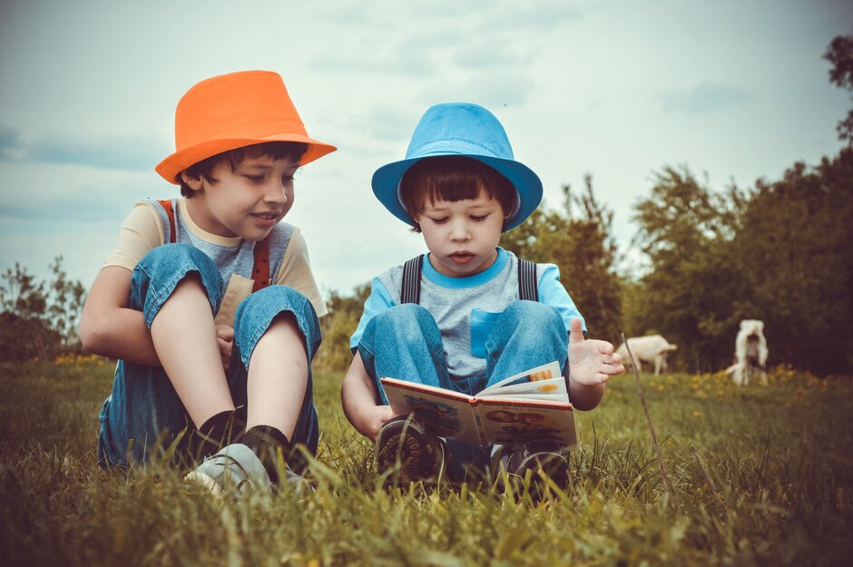 Zwei kleine Jungen mit bunten Hüten sitzen auf einem Feld und lesen ein Buch.